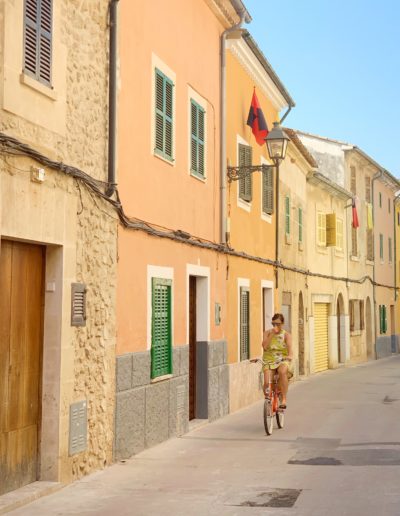 a woman rides her bike down a pastel coloured street in majorca